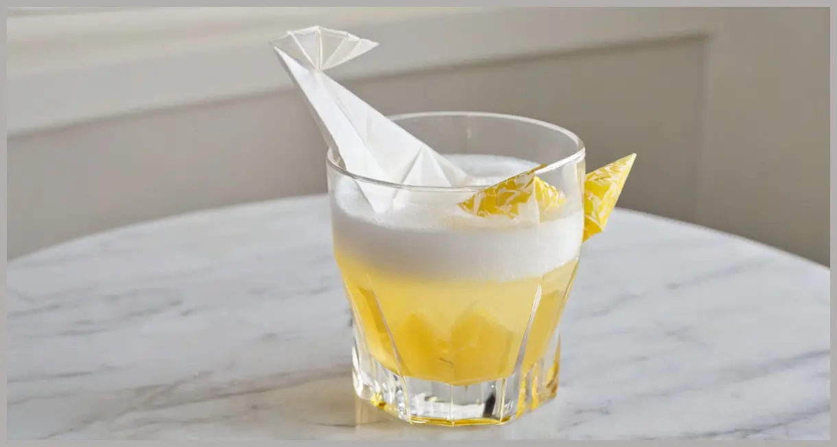 An extreme close-up, photorealistic image of a delicate origami paper boat resting on the frothy white head of an Umibozu yuzu cocktail Cocktail in a coupe glass, under soft diffused light.