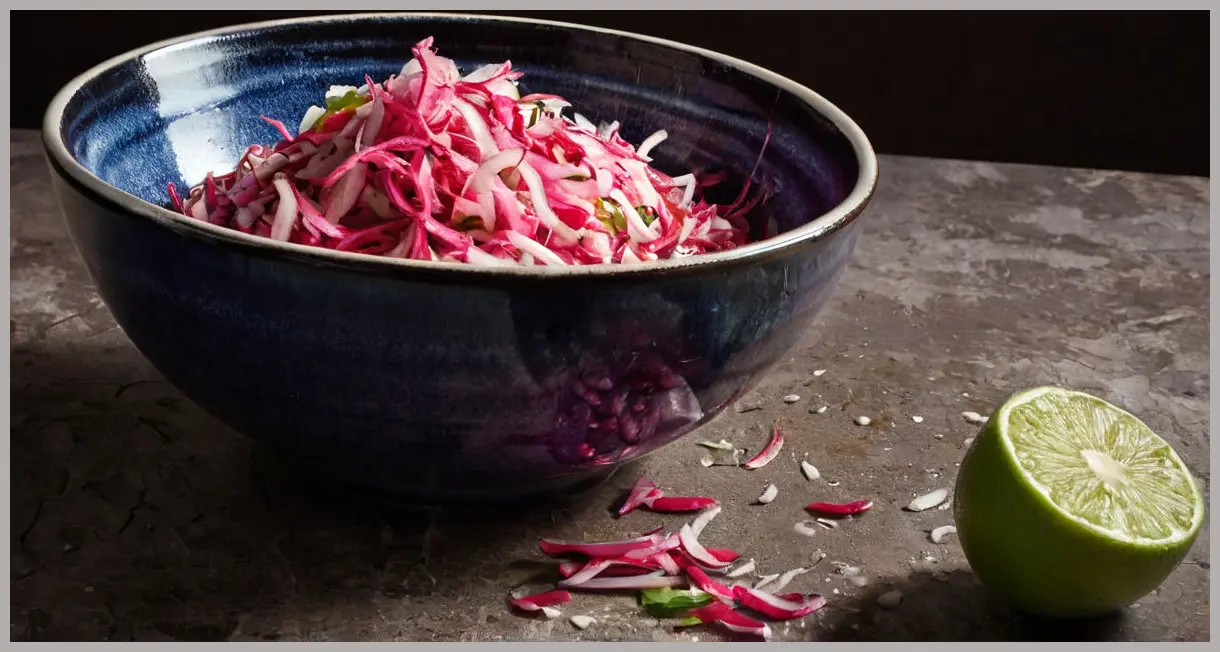 Taco slaw with pink pickled onions spilling from a ceramic bowl, dutch angle, dramatic side lighting, high contrast, moody atmosphere.