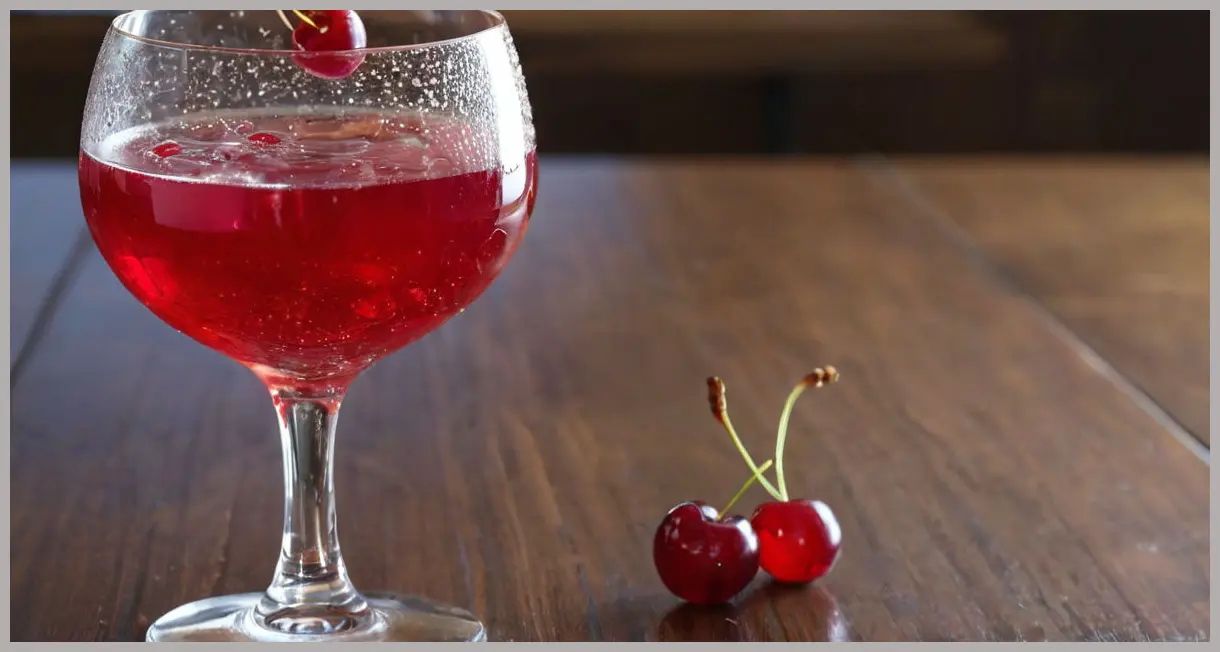 A glistening Kirsch royale cocktail in a crystal champagne flute on a dark wood bar, garnished with two vibrant red cherries on a pick.