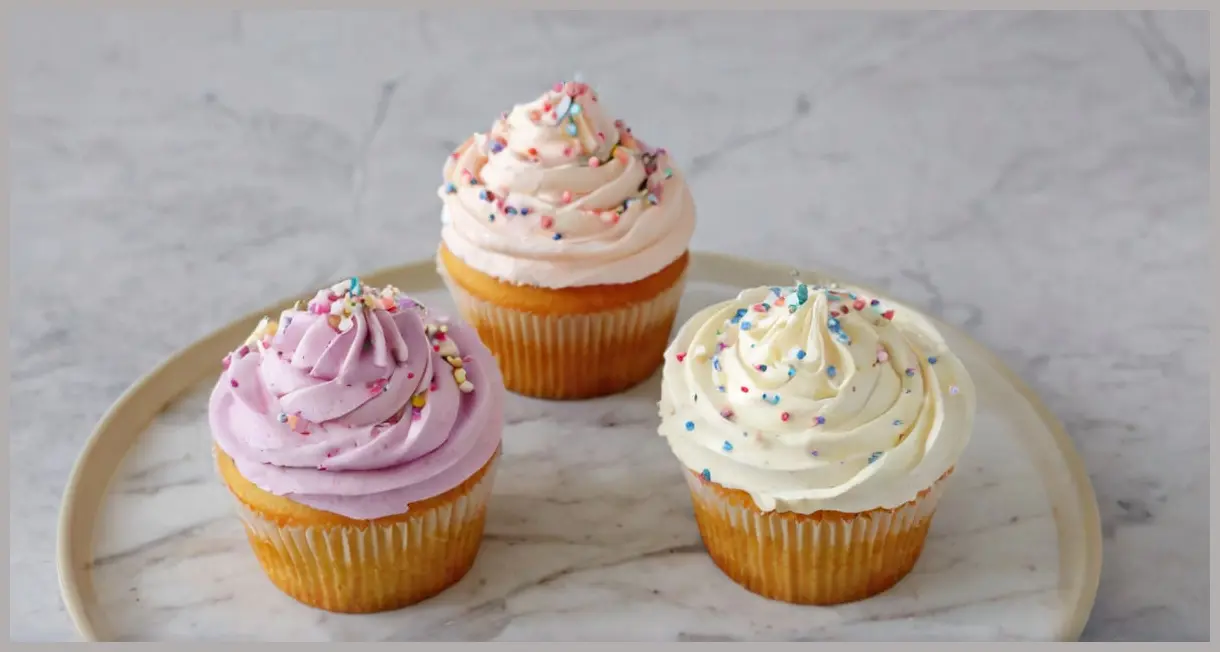Three pastel fairy cakes with minimalist buttercream piping and sprinkles, arranged on a marble surface. Simple buttercream icing