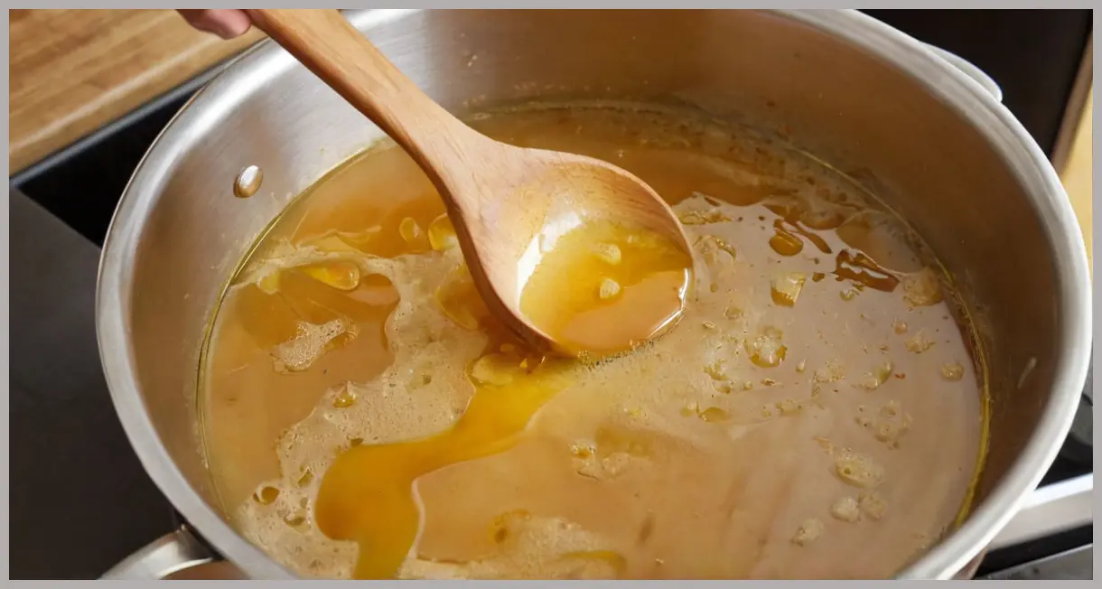 A high-angle view of hot buttered rum ingredients gently simmering in a glass saucepan, a wooden spoon stirring visible melting butter swirls and rising steam.