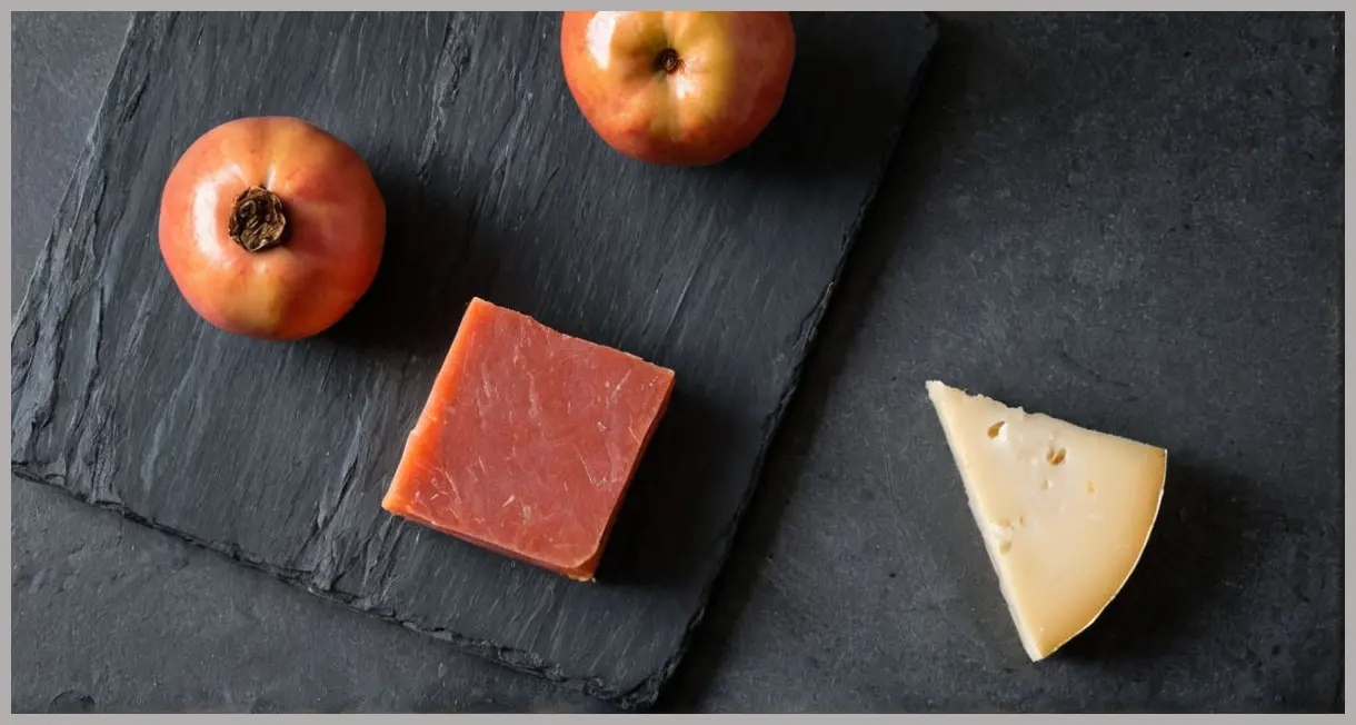 Overhead flat lay of Membrillo (quince paste) and Manchego cheese on slate, with muted earthy tones and sharp texture focus.
