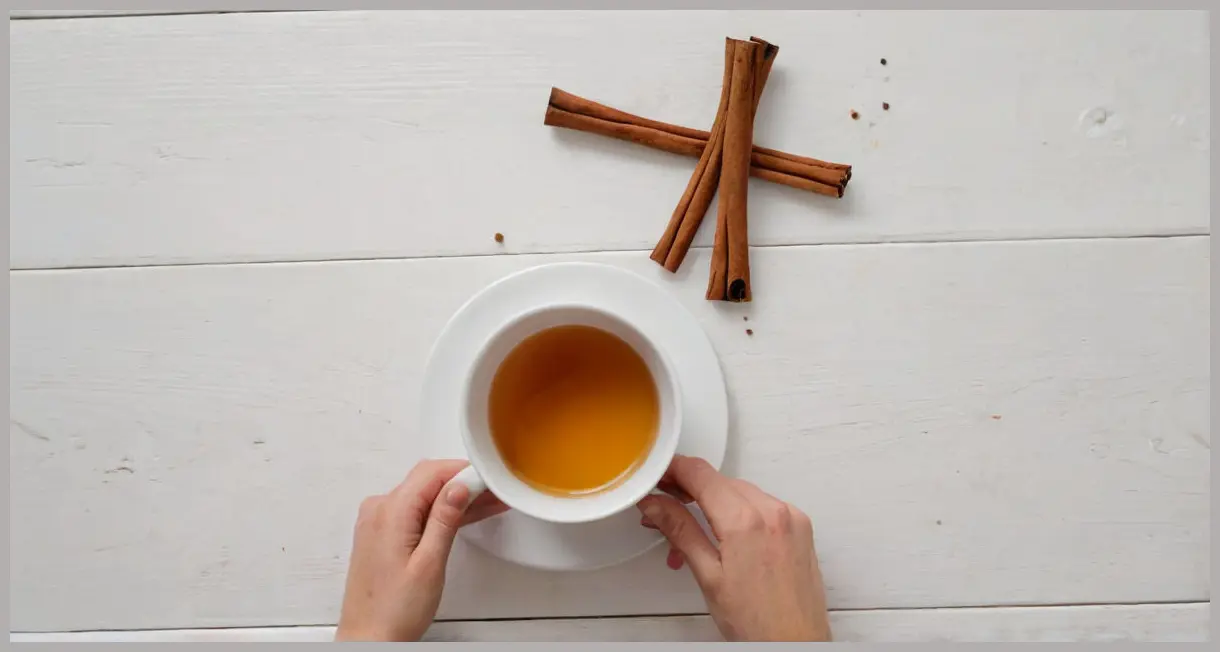 An overhead flat lay of a single hot buttered rum in a white ceramic mug, with steam, alongside whole cloves and a broken cinnamon stick on a clean white surface.