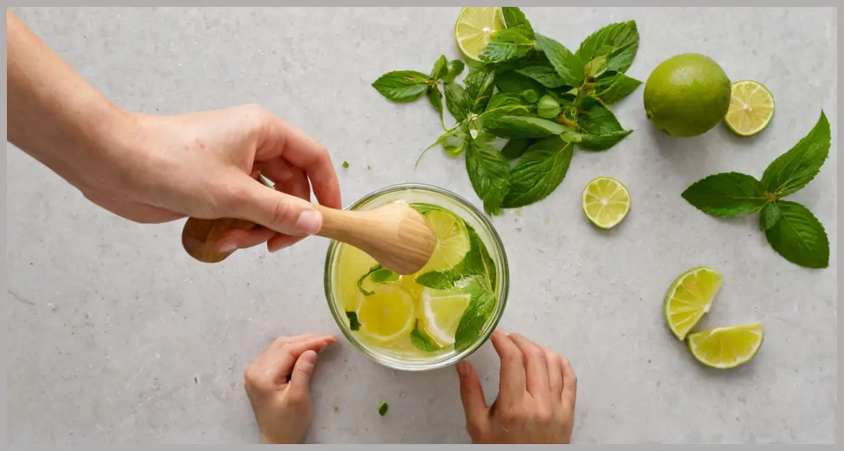 A photorealistic overhead shot showing fresh lime chunks, elderflower cordial, and mint leaves being muddled in a tall glass, forming the base of a Strawberry, lime and elderflower muddle, under soft studio lighting.