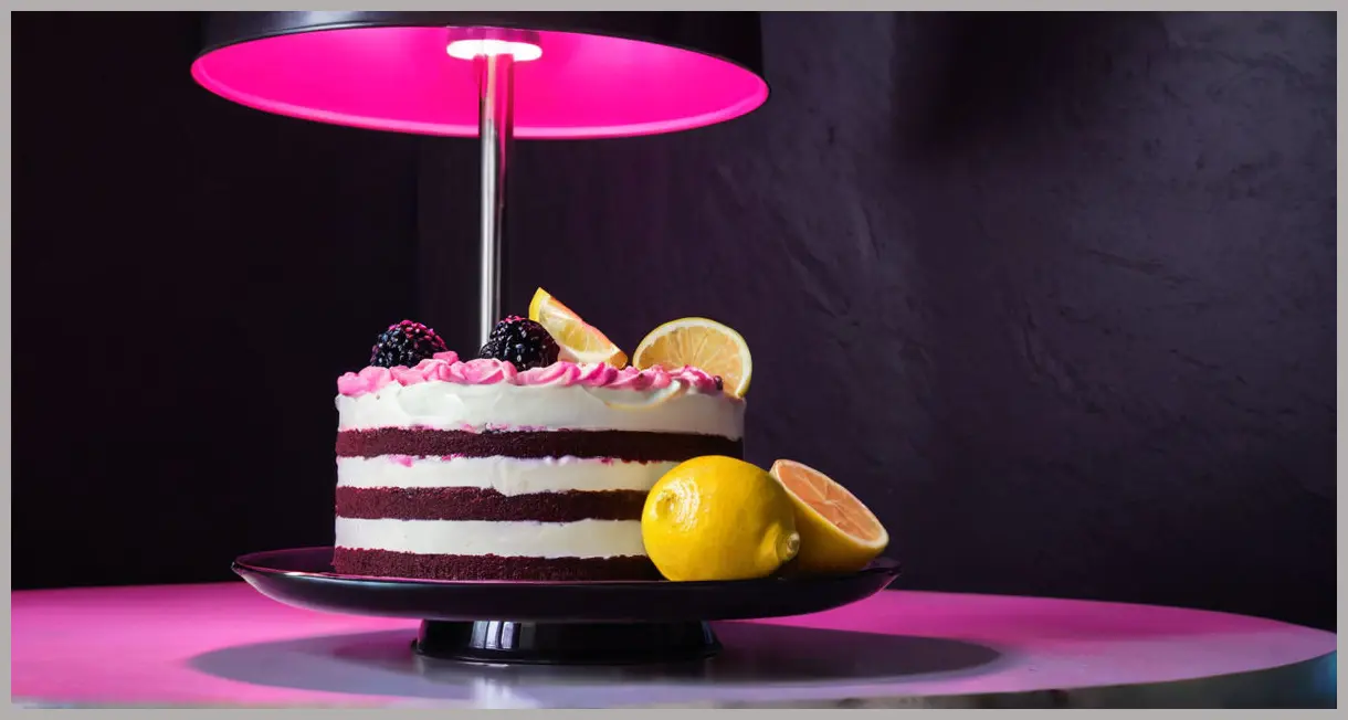 Dutch angle of a Lemon and blackberry stripe cake mid-slice under neon-pink light, knife paused in glossy frosting.