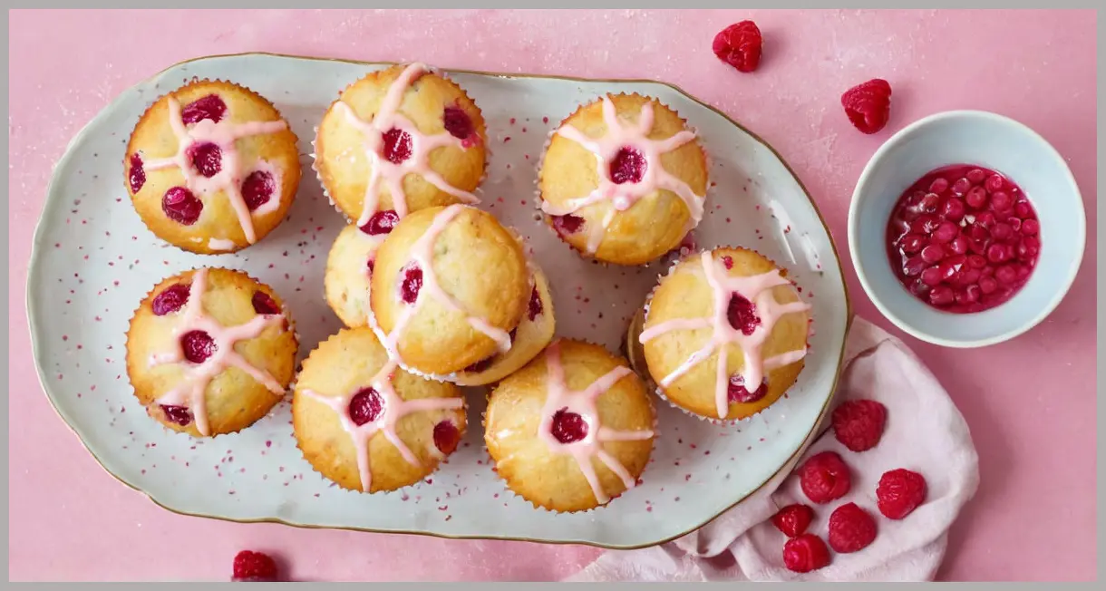 A dozen raspberry and custard muffins arranged on a vintage plate, topped with pink icing and pastel sprinkles, captured in bright natural light.