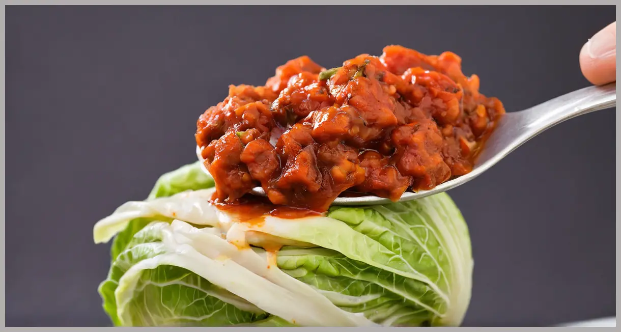 Extreme close-up of a spoonful of vegan cabbage cacciatore, layers of cabbage and olive in tomato sauce, high-contrast lighting and blurred black background.
