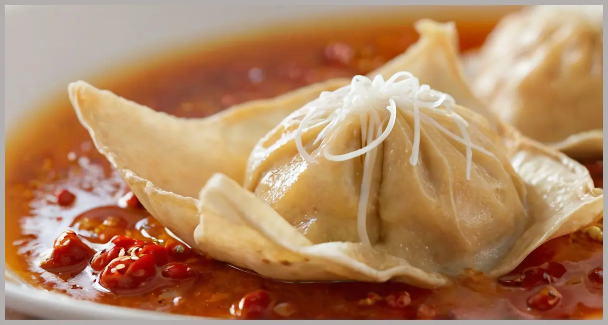 Extreme close-up of a single poached wonton glazed in chilli-garlic sauce, backlit to highlight steam and textures. Poached wontons in chilli-garlic sauce