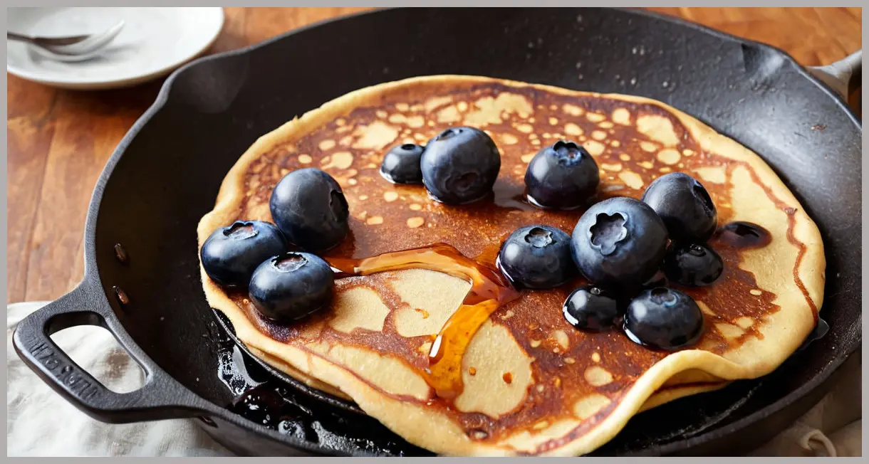 Dutch angle shot of a vegan blueberry pancake mid-flip in a cast-iron skillet, with caramelizing blueberries and sizzling syrup bubbles. Vegan blueberry pancakes