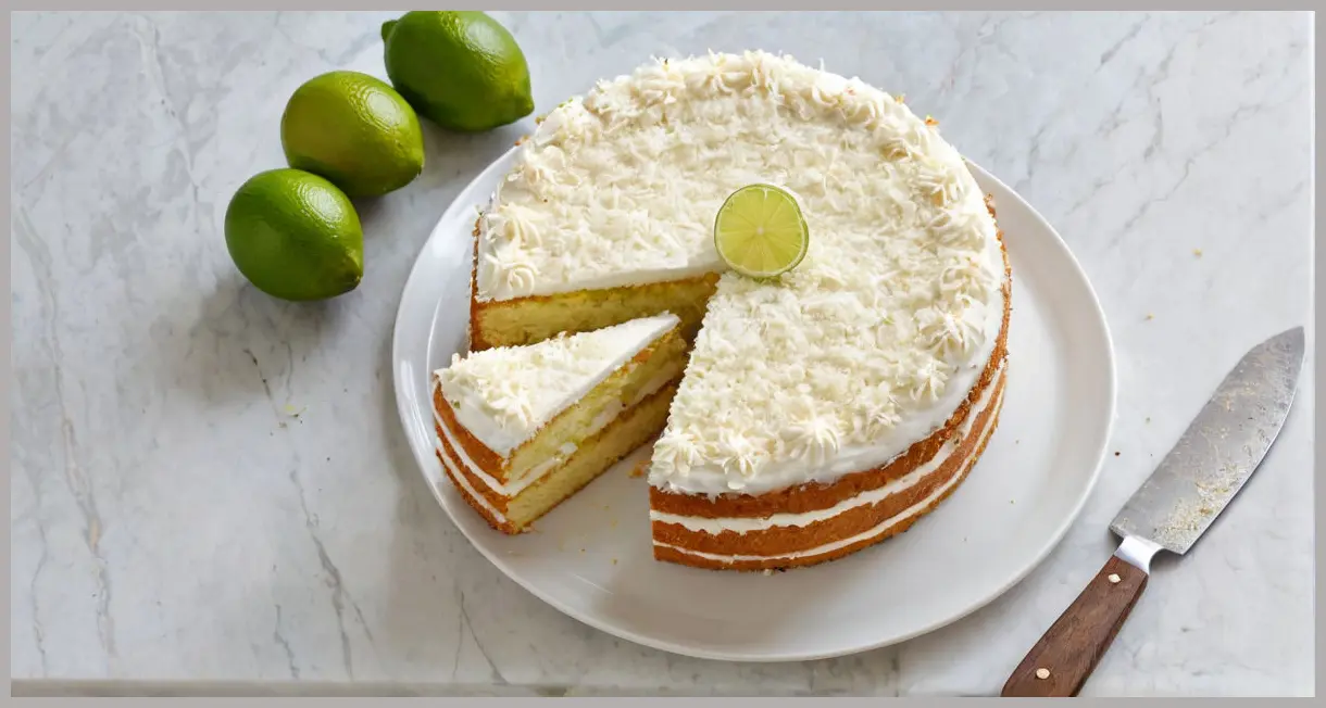 Overhead view of a sliced Coconut and lime angel cake on marble, surrounded by lime wedges and coconut flakes in bright daylight.