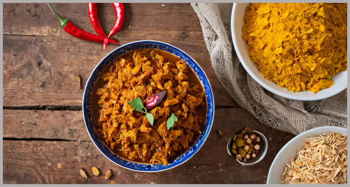 Overhead view of Jackfruit pickle (Kathal achaar) in a ceramic bowl, surrounded by scattered spices on rustic wood, soft studio light.