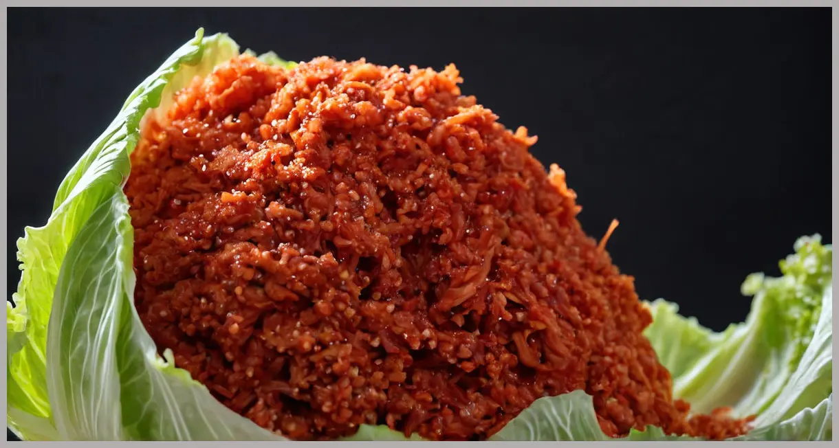 Extreme close-up of a fermented napa cabbage shred, coated in bright red gochugaru paste. Bubbles and crisp texture under dramatic light. Vegan kimchi