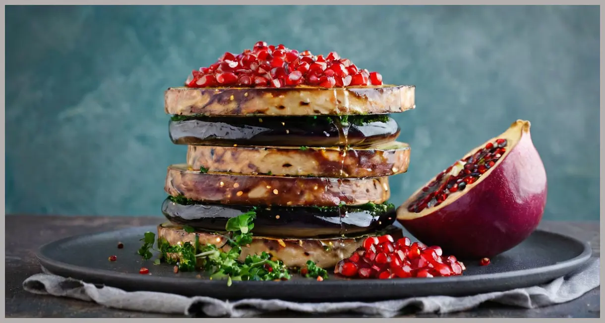 A side-angle shot of glazed aubergine slices stacked with tabbouleh spilling out, under cool blue twilight lighting. Pomegranate-glazed aubergines and courgette tabbouleh