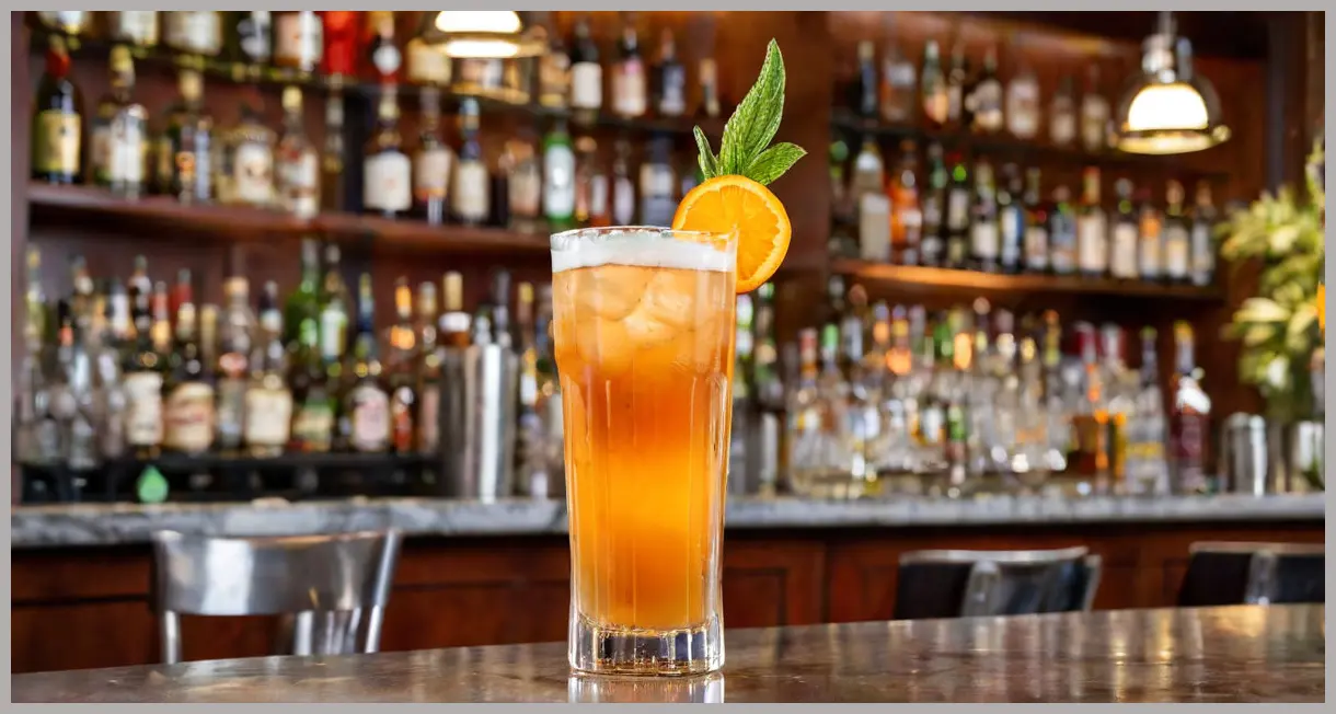 A sleek Collins glass of Pimm’s fizz and ginger ale, with tall ice spears, an orange peel spiral, and a mint leaf, in a warm bar glow.