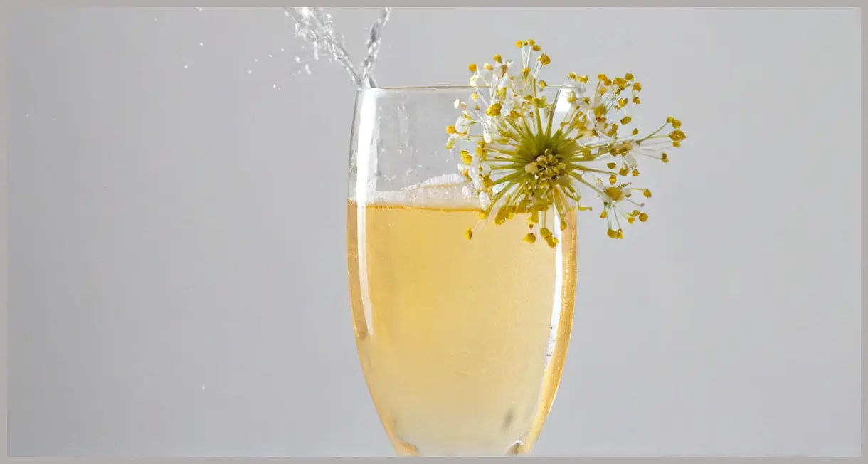 An Orchard Spritz Cocktail in a crystal champagne flute, shown mid-pour with a dramatic cascade of bubbles and foam, featuring pale elderflower cordial and an edible white flower garnish, under bright studio lighting.