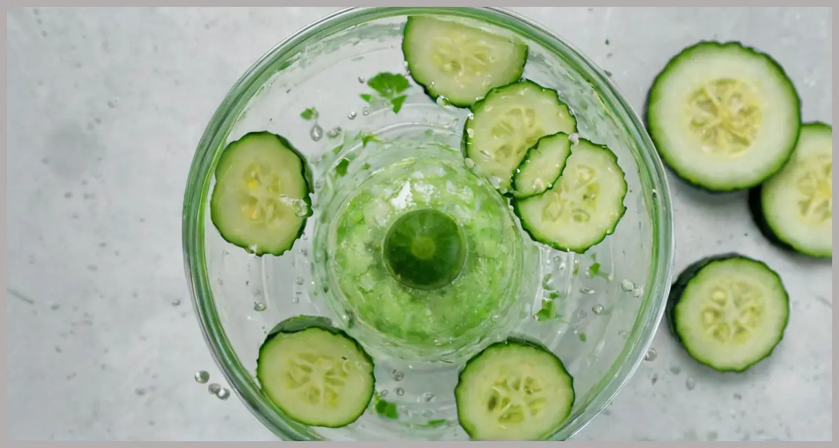 A top-down shot of a clear blender with chopped fresh cucumber, salt, and water, actively blending for a Cucumber cooler cocktail mix, under soft studio lighting.