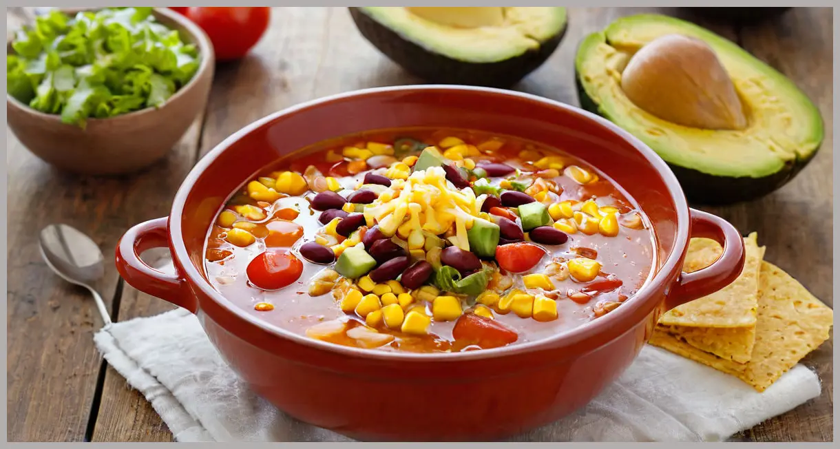 A rustic clay bowl of Mexican veggie and tortilla soup in golden hour light, topped with chips, cheese, and avocado, steam rising. Mexican veggie and tortilla soup