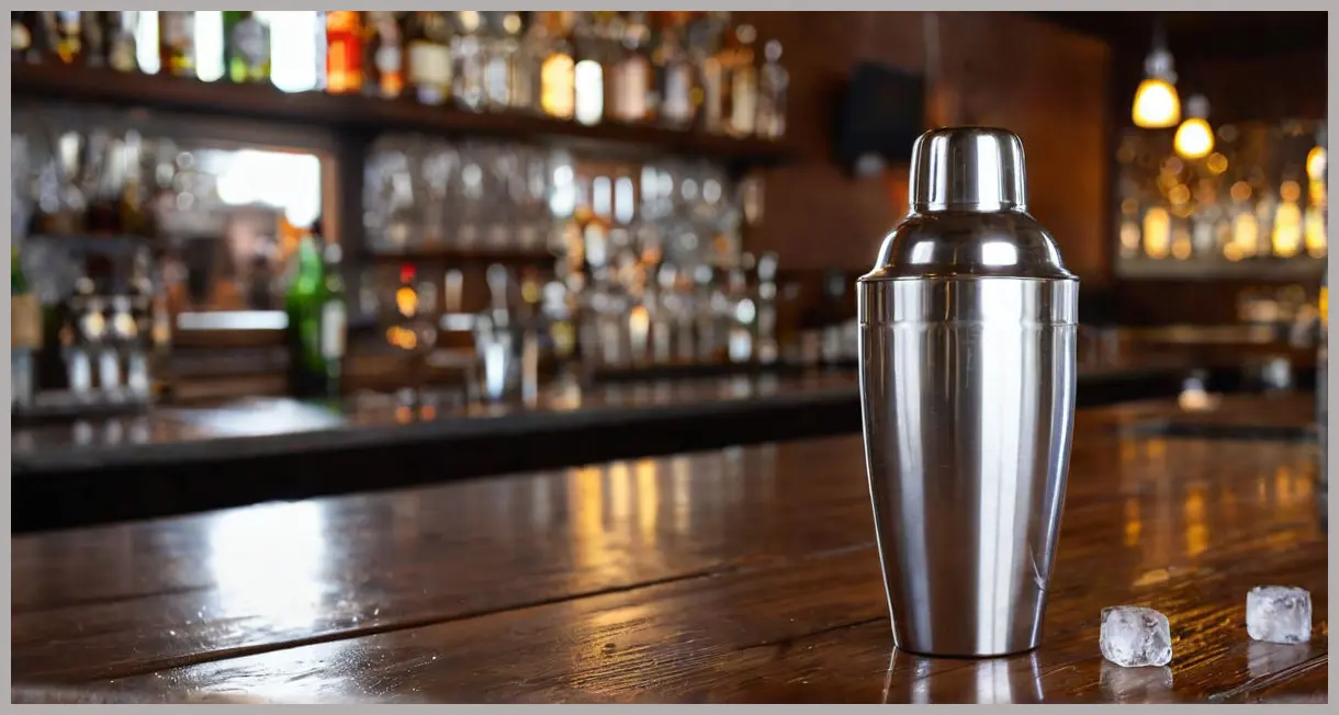 A frosty stainless steel cocktail shaker, filled with ice, showing condensation on a dark wood bar, suggesting the preparation of The Botanist Fizz.